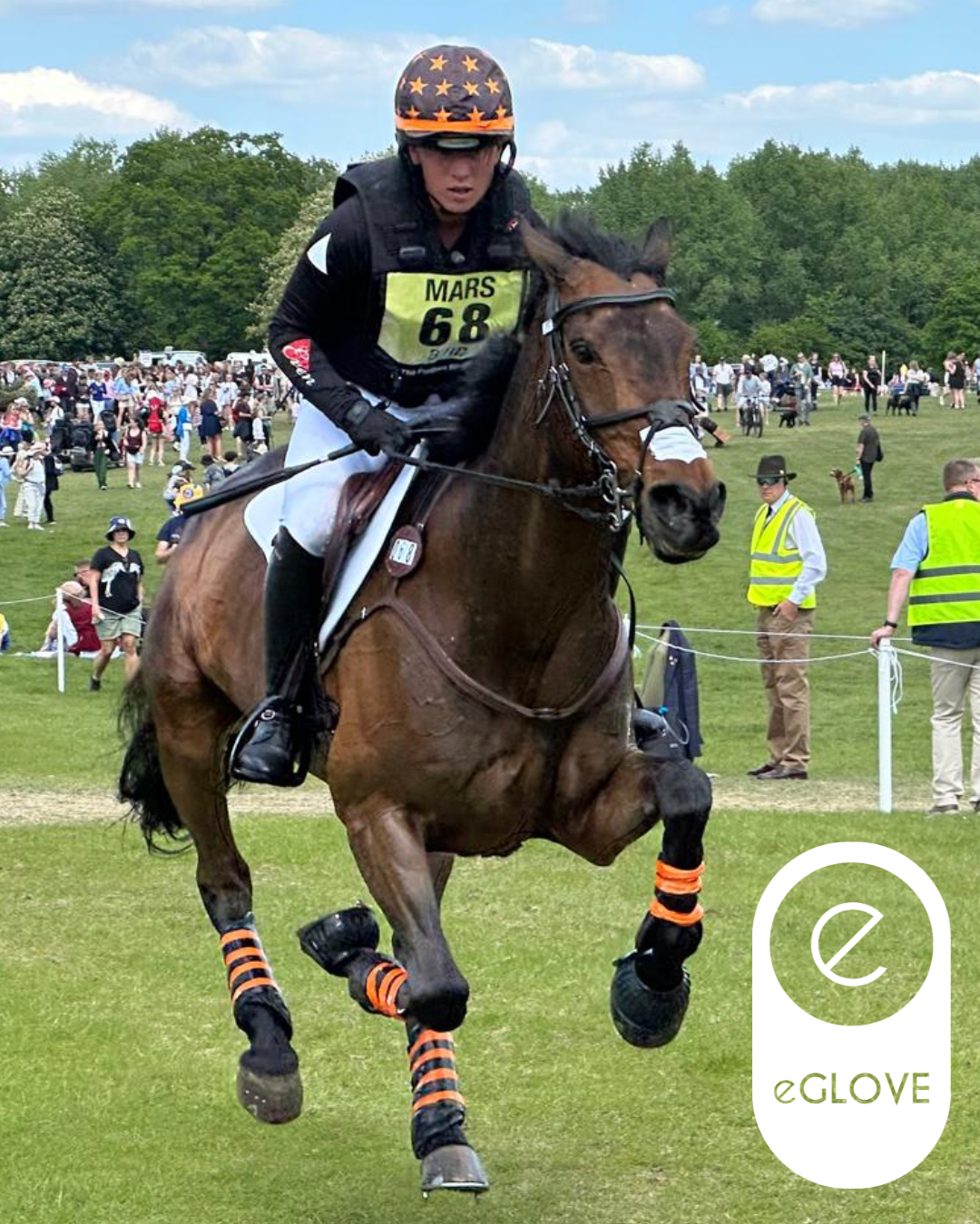 Eventing rider wearing black eGLOVE equestrian gloves competing at Mars Badminton Horse Trials on a bay horse with orange brushing boots, crowds visible in the background